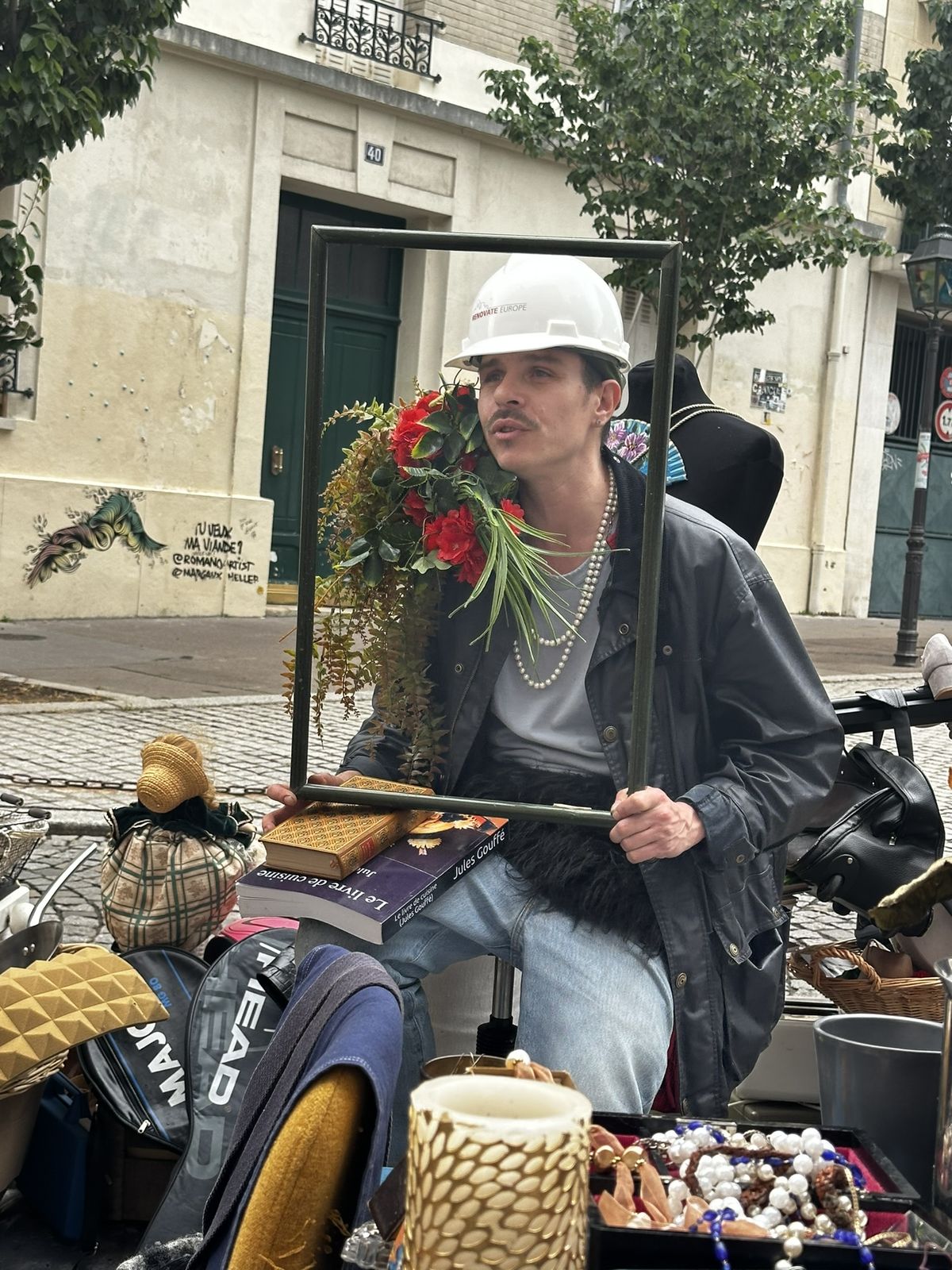 Ben tient un cadre vide devant lui, casque de chantier et fleurs, derrière un stand de vide-grenier